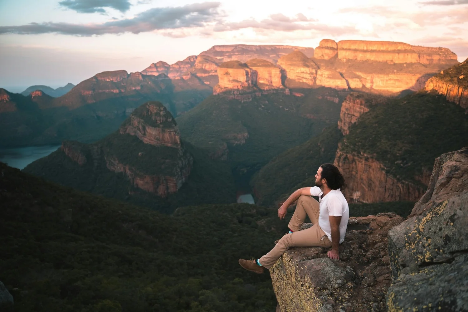 Traveller sitting on a rock ledge above a forested canyon and escarpment at sunset in Mpumalanga